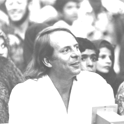 Auf dem Schwarzweißbild ist das Porträt eines Mannes zu sehen. Der Mann ist der Komponist Karlheinz Stockhausen. Er trägt ein weißes Hemd und schaut zur Seite. Im Hintergrund sind weitere Personen zu sehen.