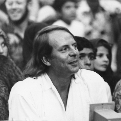 Auf dem Schwarzweißbild ist das Porträt eines Mannes zu sehen. Der Mann ist der Komponist Karlheinz Stockhausen. Er trägt ein weißes Hemd und schaut zur Seite. Im Hintergrund sind weitere Personen zu sehen.