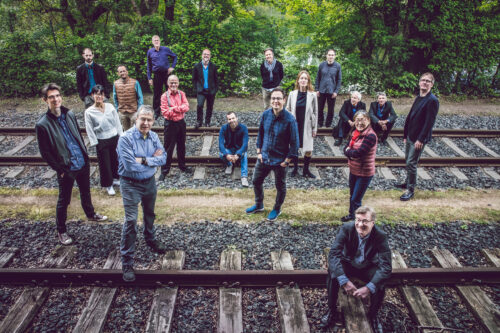Das Bild zeigt viele Menschen, die auf den Gleisen sitzen und stehen (Ensemble Modern). Im Hintergrund sieht man grüne Pflanzen.