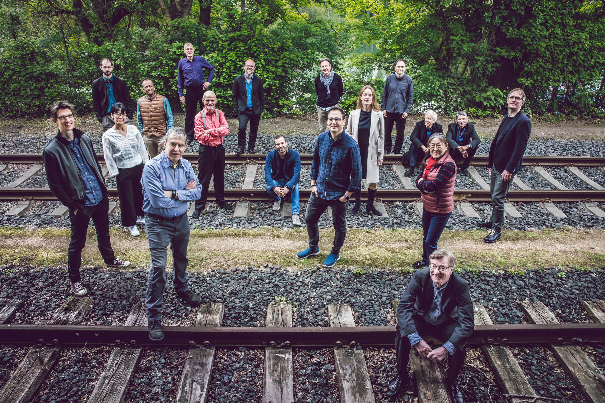 Das Bild zeigt viele Menschen, die auf den Gleisen sitzen und stehen (Ensemble Modern). Im Hintergrund sieht man grüne Pflanzen.