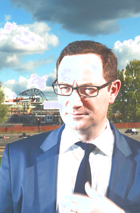 Das Porträt eines Mannes (Gordon Kampe). Er steht aufrecht und trägt einen grauen Anzug mit weißem Hemd und schwarzer Krawatte. Seine Hand ruht auf der Krawatte. Der Mann blickt weg. Im Hintergrund ist die Landschaft einer Stadt zu sehen.
