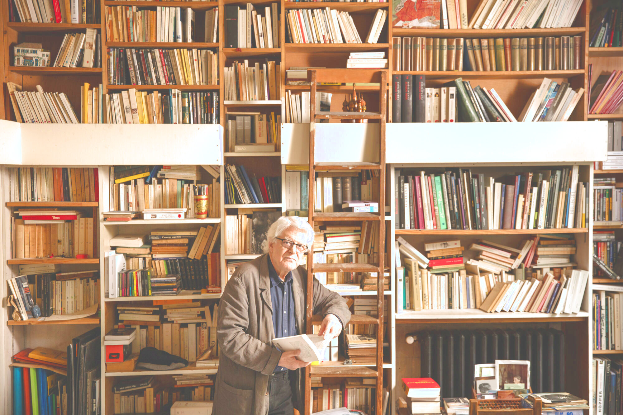 Zu sehen ist der Komponist George Aperghis. Mit einem Buch in der Hand lehnt er an einer Leiter vor einer Wand voll mit Büchern.