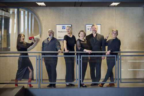 Zu sehen sind sechs Sänger:innen der Neuen Vocalsolisten in einer Ubahnstation. Die linke Person hält ein Megaphon und scheint die anderen anzuschreien, die in unterschiedlichen Posen stehen.