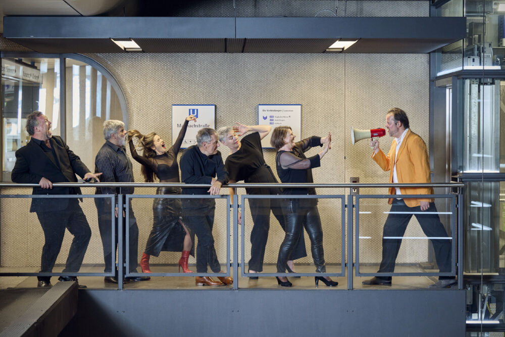 Zu sehen sind die Neuen Vocalsolisten in einer Ubahnstation. Die rechte Person hält ein Megaphon und scheint die anderen anzuschreien, die in unterschiedlichen Posen stehen.