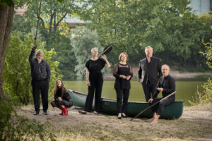 Am Flussufer stehen die sechs Sänger*innen der Neuen Vocalsolisten um ein Kanu herum.
