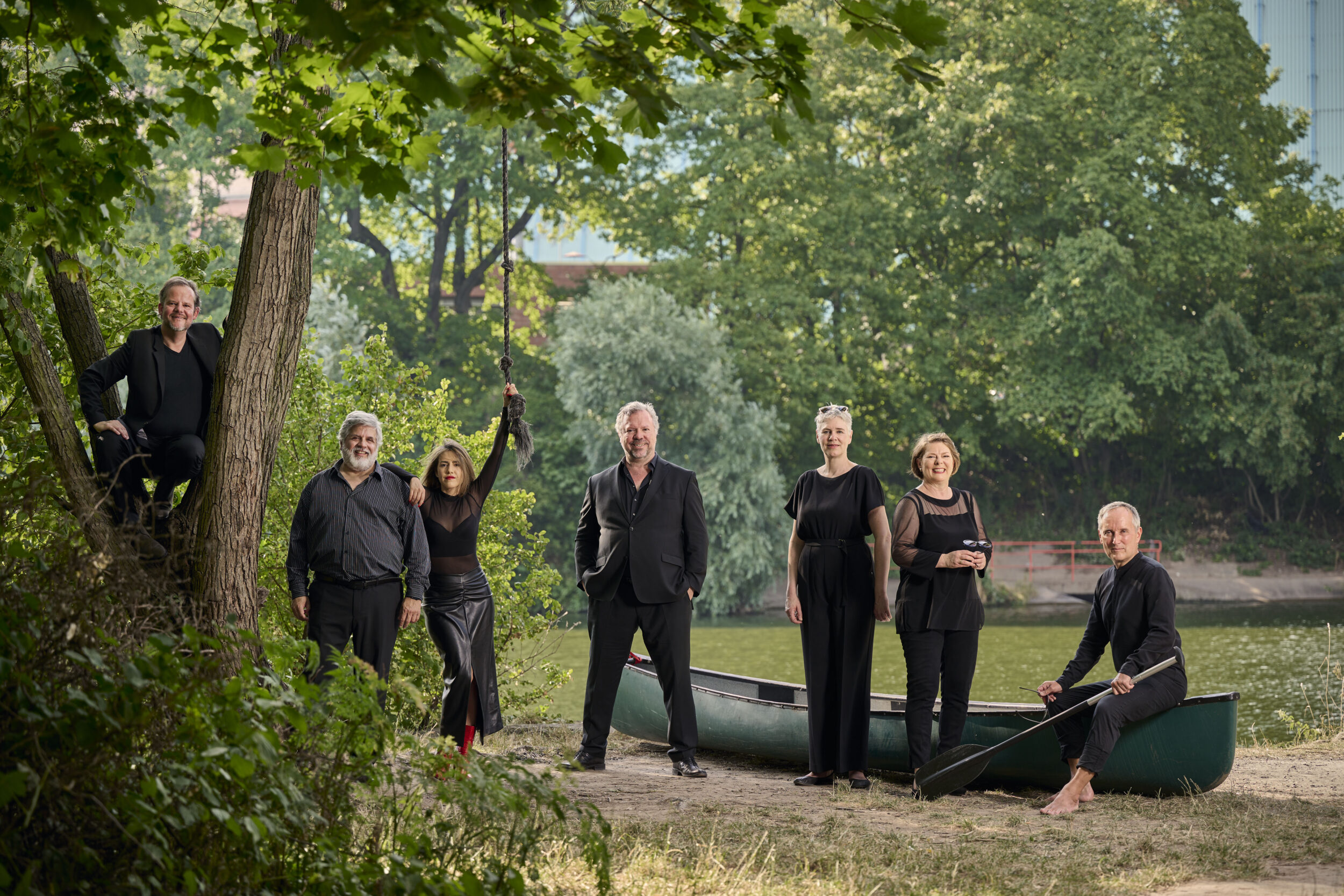 An einem Flussufer stehen die sieben Sänger:innen der Neuen Vocalsolisten. Im Hintergrund ist ein grünes Kanu zu sehen. Sie tragen alle schwarze Konzertkleidung.