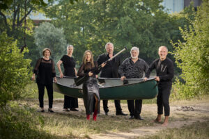 Am Flussufer stehen die sechs Sänger*innen der Neuen Vocalsolisten. Sie halten gemeinsam ein Kanu. Die Frau im Vordergrund hält ein Paddel.