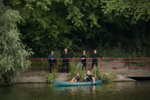 Zu sehen sind sechs Sänger*innen der Neuen Vocalsolisten. Zwei von ihnen sitzen in einem Kanu auf dem Fluss. Die restlichen vier stehen in einer Reihe am Flussufer dahinter.