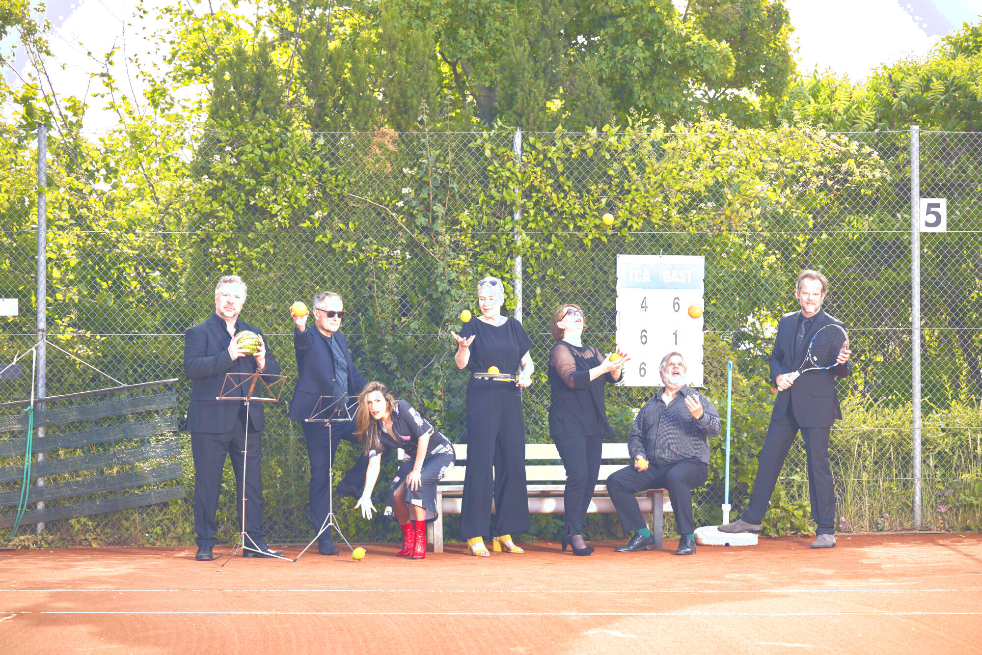 Auf dem Bild steht die Gruppe von sechs Sänger*innen der Neuen Vocalsolisten. Sie stehen auf dem Tennisplatz und halten Orangen, Zitronen, Äpfel, Melonen und Tennisschläger in den Händen.