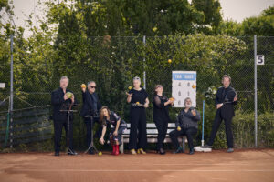 Auf dem Bild steht die Gruppe von sechs Sänger*innen der Neuen Vocalsolisten. Sie stehen auf dem Tennisplatz und halten Orangen, Zitronen, Äpfel, Melonen und Tennisschläger in den Händen.