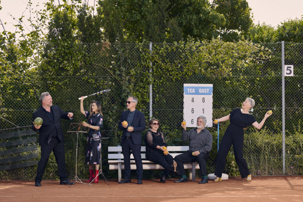 Auf dem Bild steht die sechs Sänger:innen der Neuen Vocalsolisten. Sie tragen Konezrtkleidung und. stehen auf einem Tennisplatz. Sie halten Orangen, Zitronen, Äpfel, Melonen und Tennisschläger in den Händen.