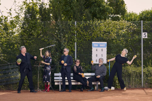 Auf dem Bild steht die sechs Sänger*innen der Neuen Vocalsolisten. Sie tragen Konezrtkleidung und. stehen auf einem Tennisplatz. Sie halten Orangen, Zitronen, Äpfel, Melonen und Tennisschläger in den Händen.