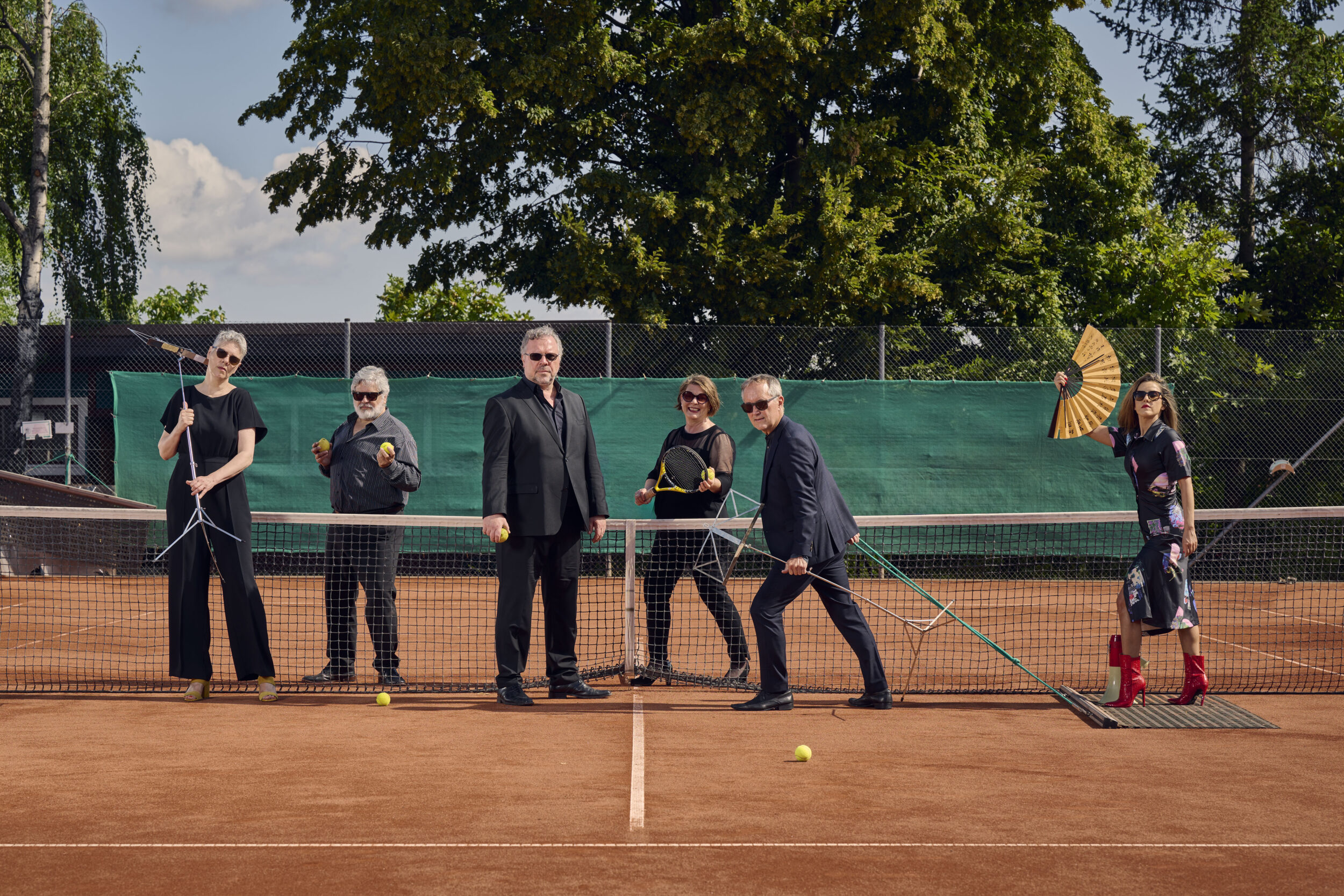 Die sechs Sanger*innen der Neuen Vocalsolisten befinden sich auf einem Tennisplatz. Sie tragen Konzertkleidung. Ein Mann zieht eine Frau auf dem Schleppnetz. Sie hält einen Fächer. Die anderen halten Tennisbälle und Schläger sowie Notenständer in ihren Händen.