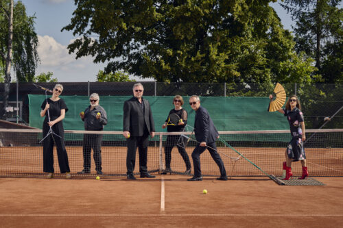 Die sechs Sanger*innen der Neuen Vocalsolisten befinden sich auf einem Tennisplatz. Sie tragen Konzertkleidung. Ein Mann zieht eine Frau auf dem Schleppnetz. Sie hält einen Fächer. Die anderen halten Tennisbälle und Schläger sowie Notenständer in ihren Händen.