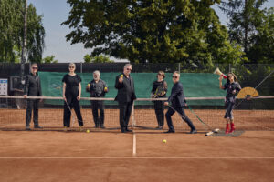 Die sieben Sänger*innen der Neuen Vocalsolisten befinden sich auf einem Tennisplatz. Sie tragen schwarze Kleidung. Ein Mann zieht eine Frau auf dem Schleppnetz. Sie hält ein Megaphon und einen Fächer. Die anderen halten Tennisbälle und Schläger sowie Notenständer in ihren Händen.
