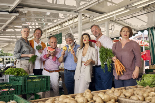 Zu sehen sind die sieben Sänger:innen der Neuen Vocalsolisten. Sie stehen inmitten eines Marktstandes. Jede:r Sänger:in hält ein Stück Obst oder Gemüse in der Hand.