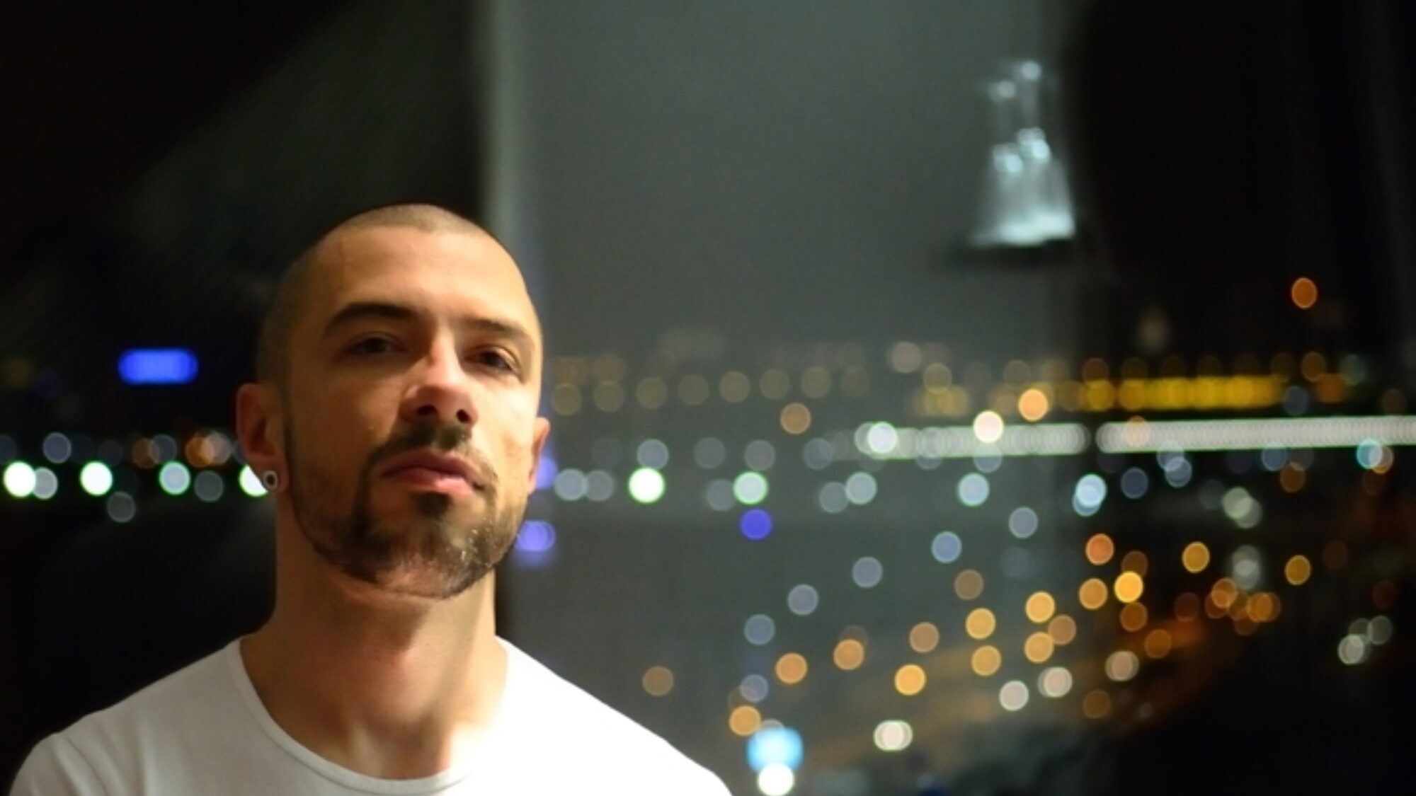 Das Porträt eines Mannes (Marko Nikodijević). Er blickt geradeaus und trägt ein weißes T-Shirt. Im Hintergrund sind die Lichter einer nächtlichen Stadt zu sehen.