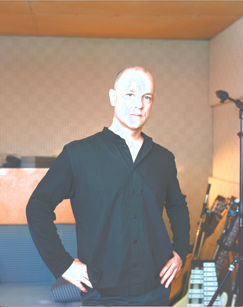 Porträt eines Mannes (Marcus Schmickler). Er steht aufrecht, die Arme an den Seiten. Er trägt ein schwarzes Hemd. Der Mann befindet sich in einem Proberaum mit Musikinstrumenten.