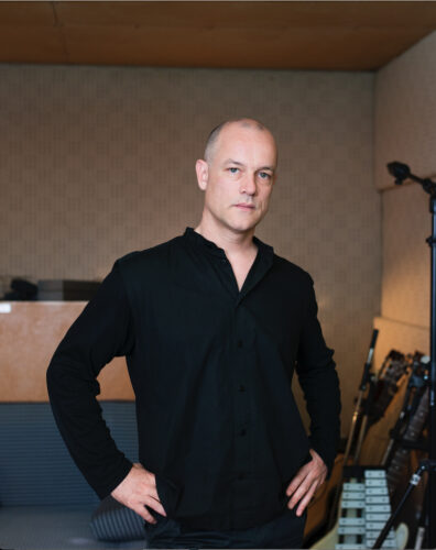 Porträt eines Mannes (Marcus Schmickler). Er steht aufrecht, die Arme an den Seiten. Er trägt ein schwarzes Hemd. Der Mann befindet sich in einem Proberaum mit Musikinstrumenten.