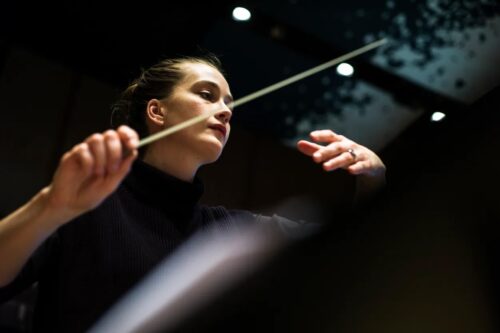 Das Bild zeigt die Dirigentin Elena Schwarz während des Dirigierens. Sie wurde von unten fotographiert. In ihrer Hand hält sie einen DIrigierstab.