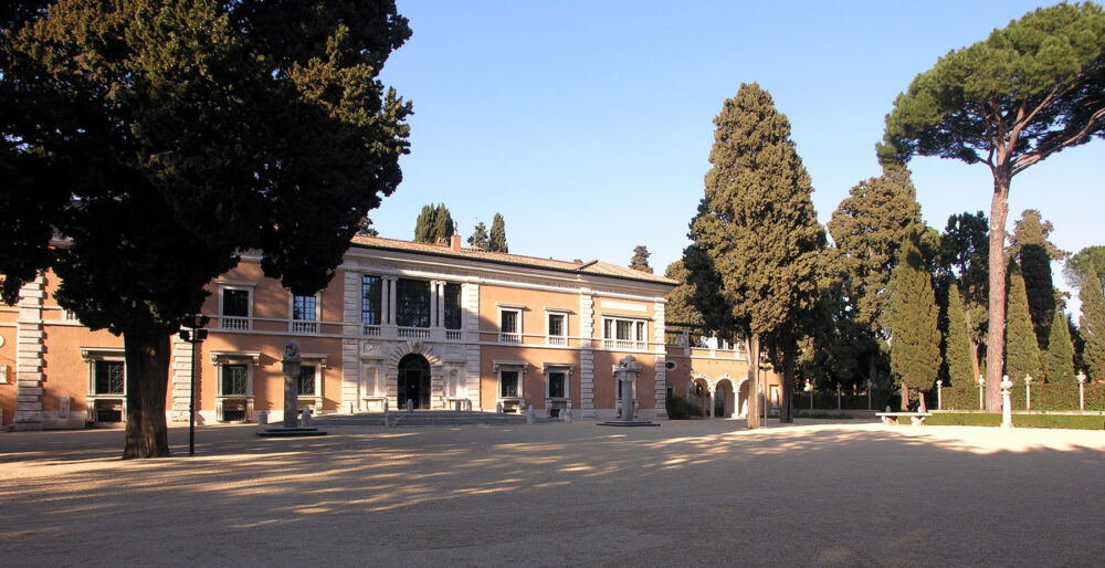 Das Bild zeigt das Gebäude der Villa Massimo. Es hat eine gelblich-rosa Farbe mit weißen dekorativen Details. Die Villa liegt in einem Garten mit südländischen Pflanzen.