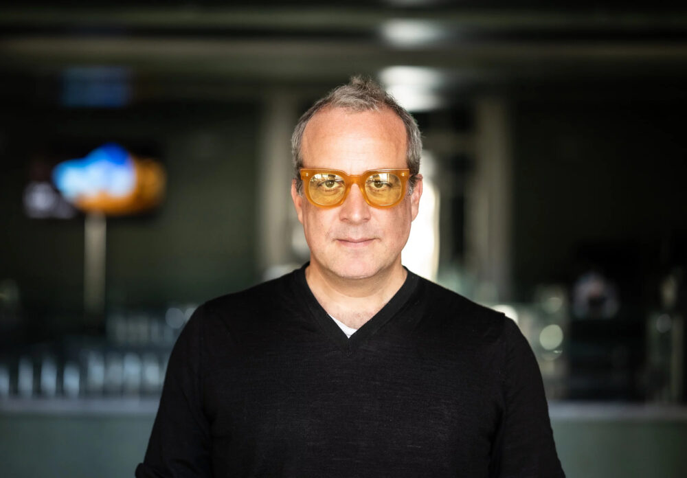 Zu sehen ist der Komponist Marc Sinan. Er trägt ein schwarzes Oberteil und eine orangefarbene getönte Brille.