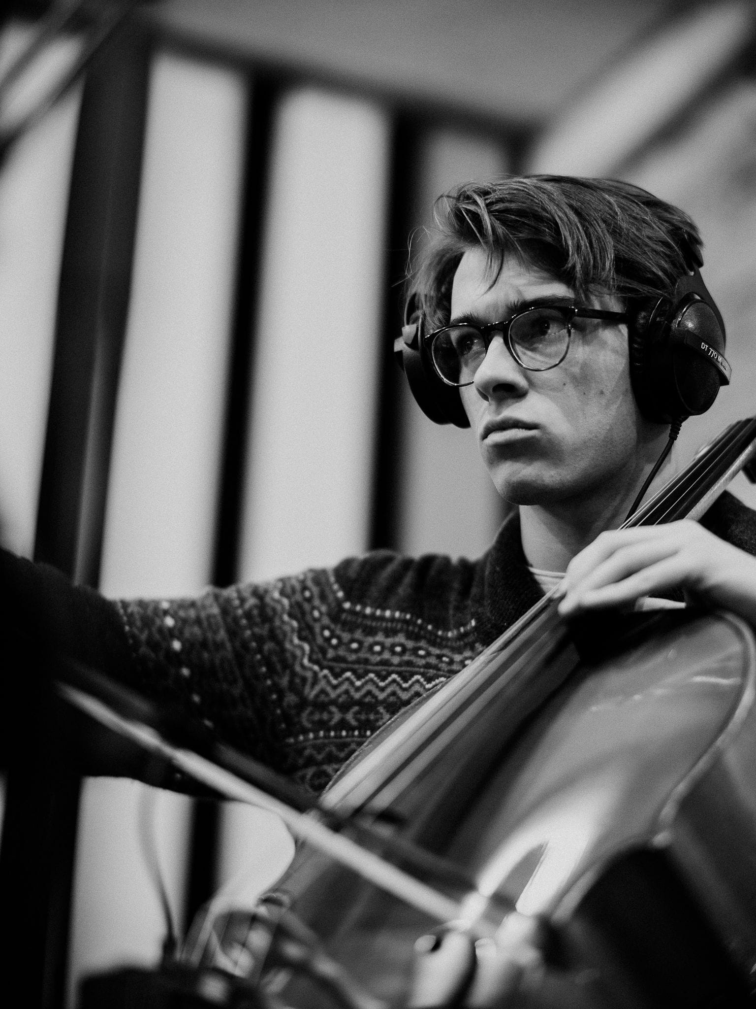 Das schwarz-weiß Foto zeigt Jakob Riecke. Er spielt konzentriert Cello und trägt dabei Kopfhörer. Er ist in einen gemusterten Pullover gekleidet, trägt eine Brille und hält den Cellobogen in der rechten Hand. Mit der linken hält er sein Cello.