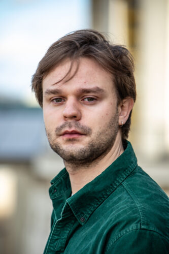Zu sehen ist Johannes Burgert mit braunen, leicht zerzausten Haaren und einem Bart. Er trägt ein dunkelgrünes Cordhend, hat einen kleinen Ohrring und blickt ernst in die Kamera. Der Hintergrund ist unscharf und zeigt helle, neutrale Farbtöne, die auf eine städtische Umgebung hindeuten.