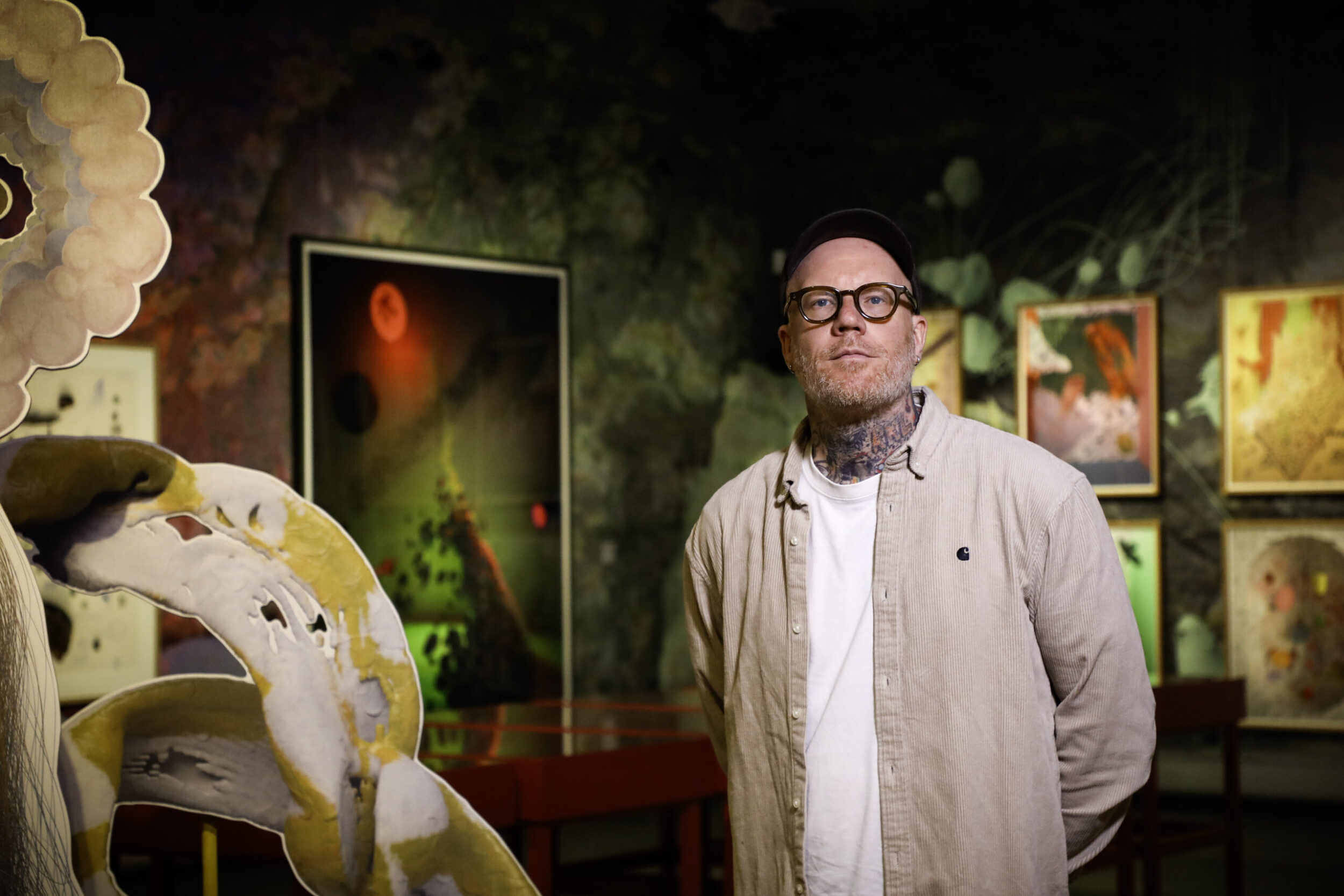 Zu sehen ist Tim Berresheim mit Brille und Halstätowierung. Er steht in einem Kunstraum oder einer Galerie und trägt ein helles, beiges Hemd. Im Vordergrund links ist eine große, abstrakte Skulptur in gelben und weißen Farbtönen zu sehen. Im Hintergrund hängen mehrere gerahmte Kunstwerke an der Wand.